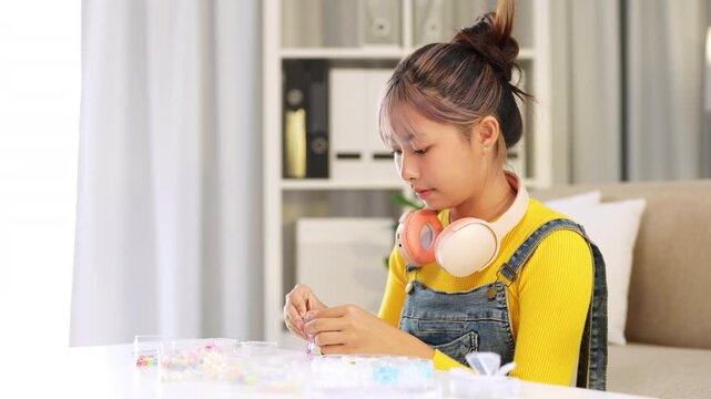Concentrated girl making bracelets with colorful beads, sitting on a sofa in the living room, entertaining herself with a creative hobby enjoying her free time in her living room.