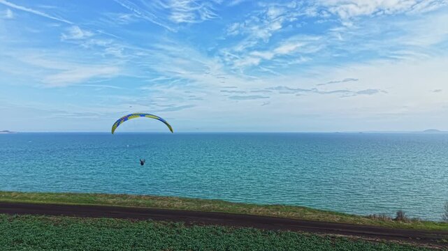 A person is paragliding above the blue water close to a grassy shore. The sky is clear and filled with clouds on a bright day, and a road runs alongside the coast.