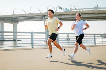 Fototapeta premium Joyful couple man and woman enjoying a morning run along the waterfront pathway under a clear blue sky
