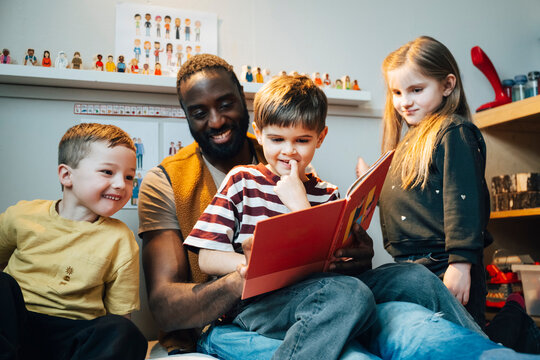 Smiling male teacher spending leisure time while reading book with students in classroom