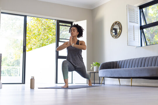 Woman practicing low lunge on yoga mat in living room wearing sportswear near water bottle