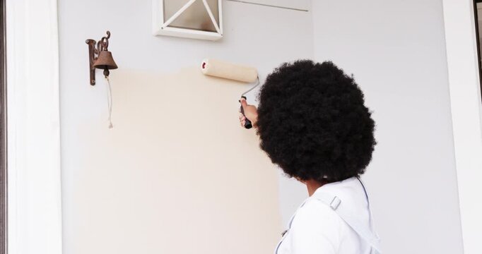 African woman placing roller under vent, repainting entryway wall near bell with tray, copy space