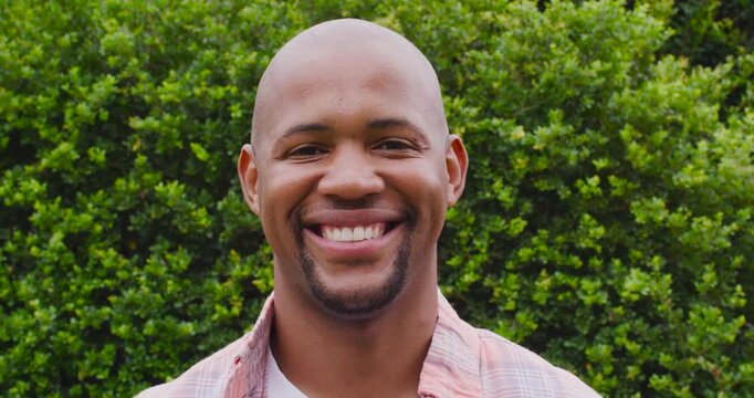 Adult man starting grin, smiling, laughing, leaning by green hedge in plaid shirt over white tee