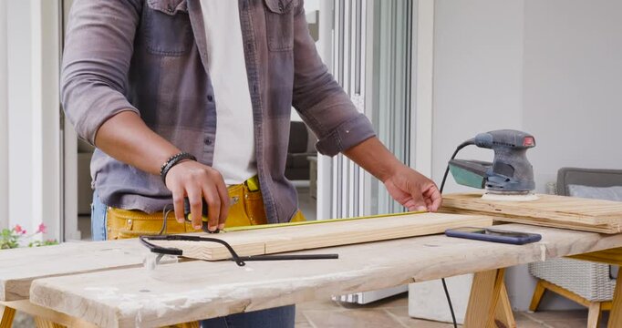 Man extending tape measure and marking wooden plank on porch bench, preparing orbital sander