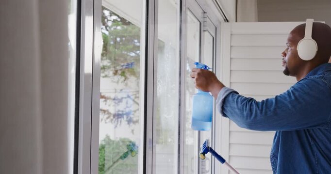 African American man spraying blue bottle and squeegeeing glass doors at home to clean, copy space