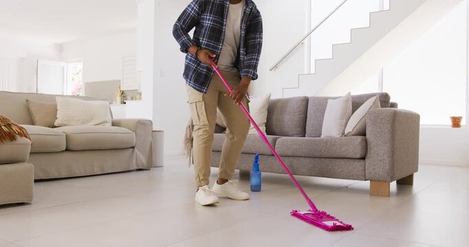 African American man picking up bottle then mopping to clean living room floor with pink mop