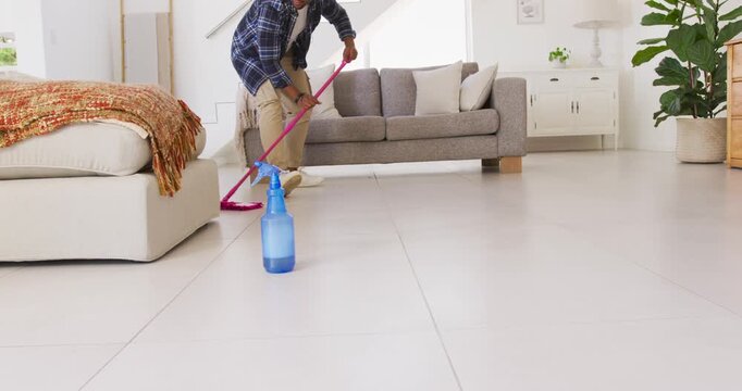 Adult African American man mopping floor in living room with pink mop after spraying blue bottle