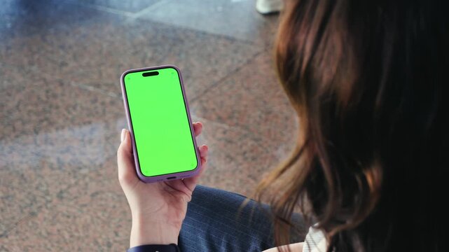A woman sits in an airport terminal, using her phone, watching movies, reading the news, spending time on social media while waiting her flight. Phone with a green screen.