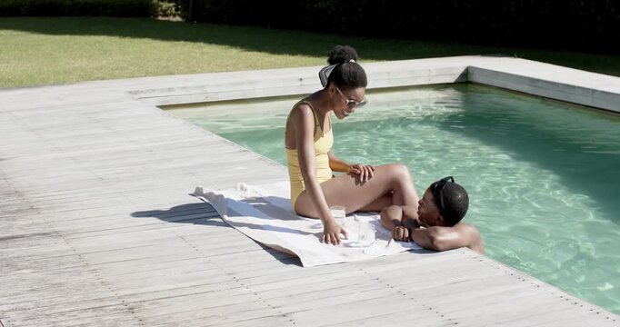 African American couple chatting by pool, woman in yellow suit reaching glass on towel, copy space