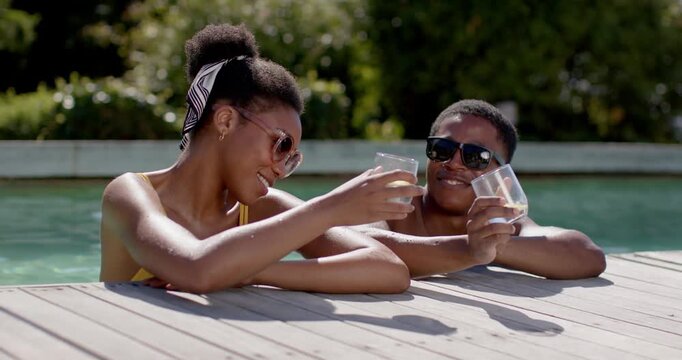 African American couple glancing, turning toward each other at pool edge, clinking glasses toasting