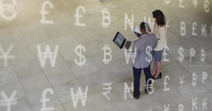Two colleagues opening laptops, reviewing finance while currency symbols floating around screens