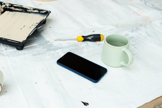Work area showing smartphone with blue case lying on drop cloth near mug screwdriver paint tray