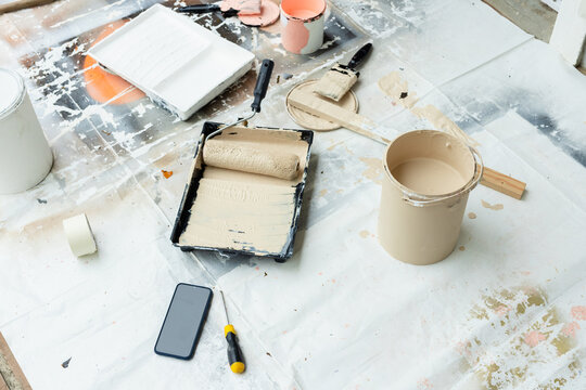 Painting area on drop cloth showing roller in tray with beige can, brush, tape phone screwdriver