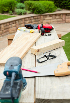 Backyard woodworking bench showing orbital sander, handsaw, safety glasses, earmuffs, tape