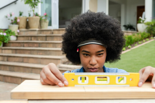 African American woman aligning yellow spirit level on wooden boards at porch wearing denim shirt