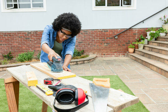 Adult African woman sanding wood on home patio with orbital sander wearing denim jacket and goggles