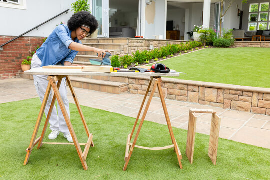 African American woman using sander on plank on lawn patio, wearing denim shirt, copy space