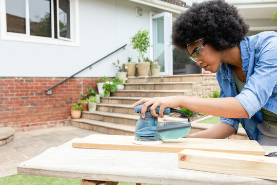 African American woman sanding wooden plank using orbital sander on front patio wearing goggles