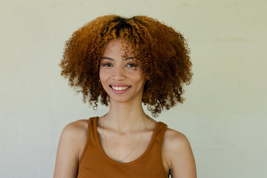 African woman smiling, posing in front of beige wall wearing rust top, silver necklace and studs