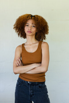 African American woman standing with arms crossed against light wall in brown tank top, dark jeans