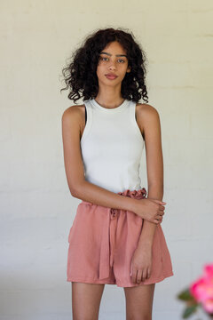 African American woman posing against pale wall, wearing white top, salmon shorts near pink flowers