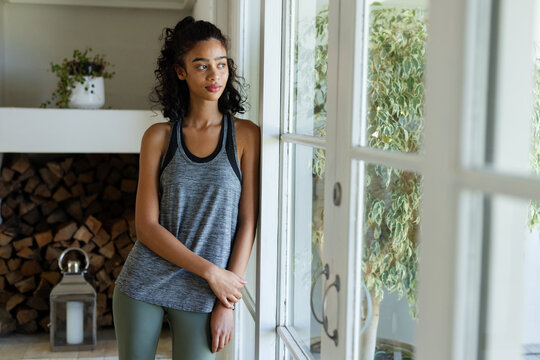 African American woman leaning on frame in grey tank, olive leggings gazing through glass at home