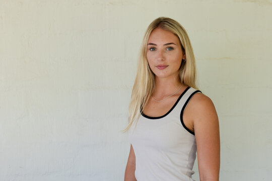 Woman standing by cream plaster wall wearing white tank with black trim, thin necklace, copy space
