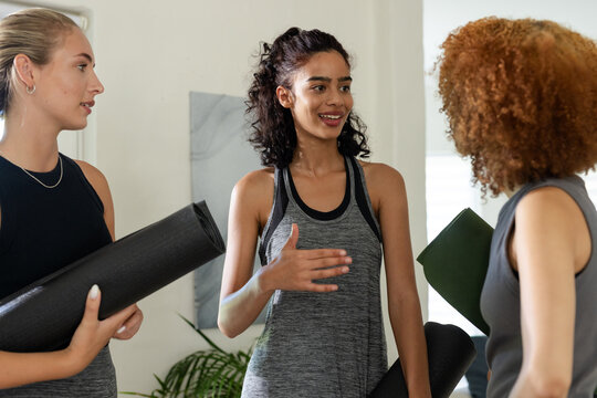 Female friends standing in yoga studio holding rolled mats in tanks, chatting near bright window