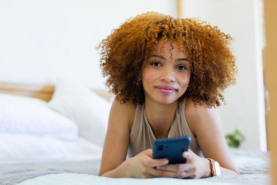 African American woman lying on bed in bedroom holding smartphone and wearing sleeveless beige top