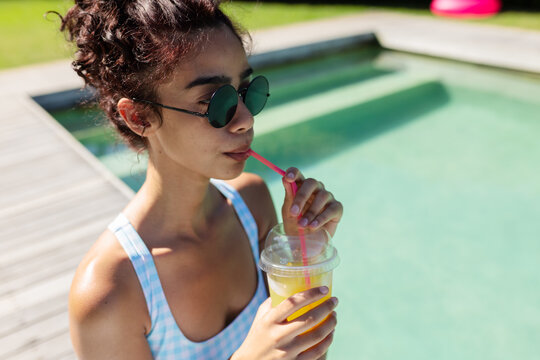African American woman standing on deck by pool, sipping from plastic cup, in gingham swimsuit