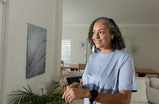 Middle-aged African woman tapping black smartwatch while wearing white earbuds in living room