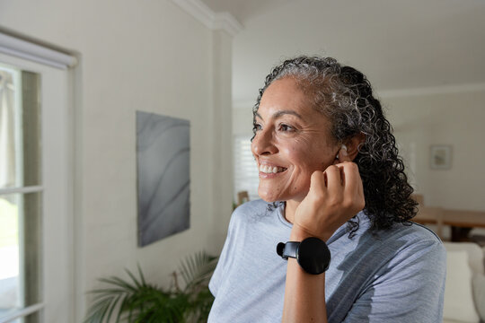 African American woman adjusting earbud while smiling by window at home, showing black smartwatch