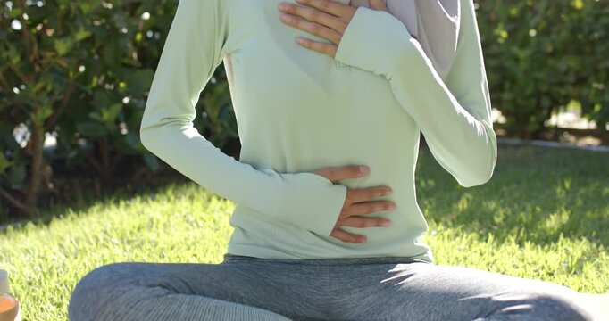 African American woman placing hands on abdomen, breathing calmly on yoga mat sunlit lawn in hijab