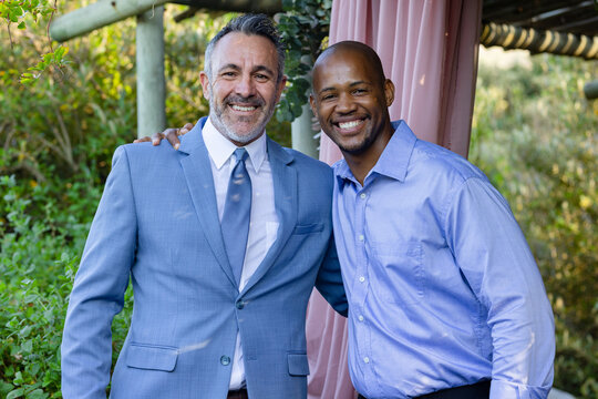 Diverse male friends standing smiling under pergola draped pink drapery, wearing suit jacket