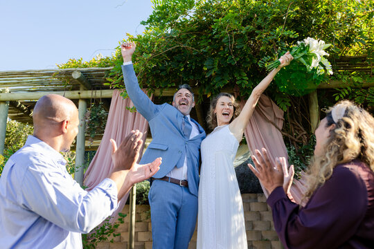 Newlywed couple raising white lily bouquet and cheering with guests clapping under pink pergola
