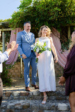 Couple bride in white lace groom in suit descending steps beneath vine pergola holding bouquet