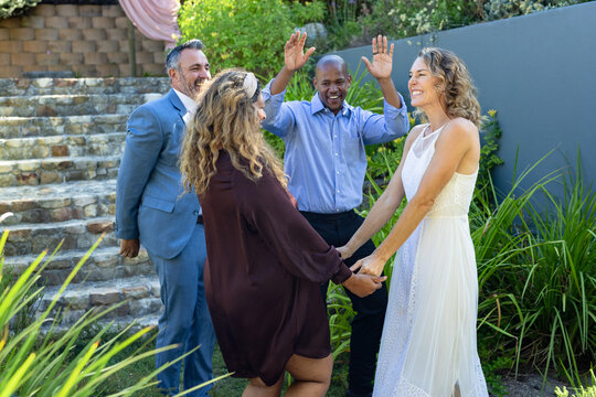 Four friends holding hands and celebrating in backyard garden, woman in white lace dress near steps