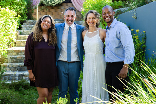Two couples standing together in backyard garden before draped fabric arch wearing suit and dress