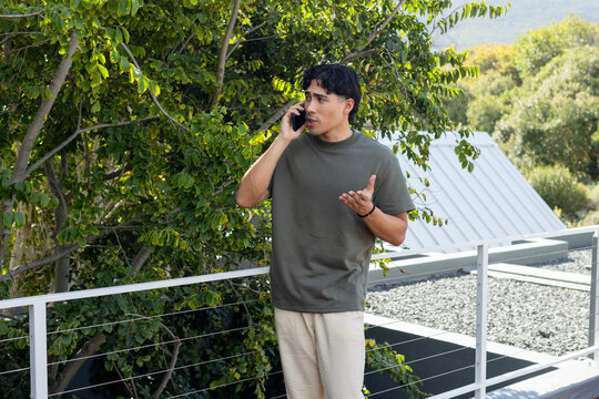 Asian man in 20s talking on smartphone and gesturing on balcony with metal railing, olive shirt