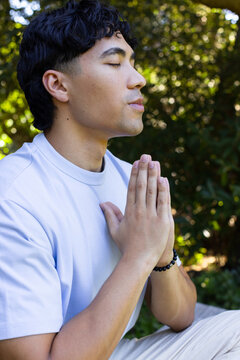 Asian man sitting in garden praying with folded hands wearing light blue tee and beaded bracelet