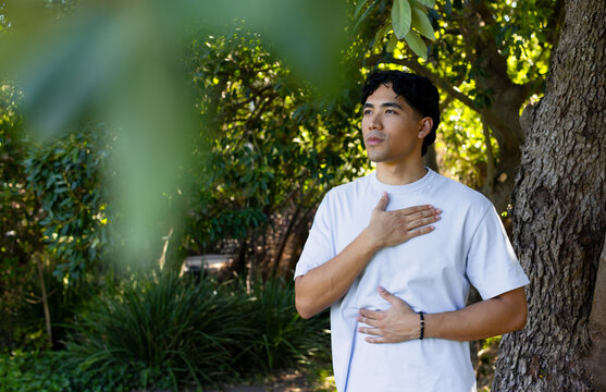 Asian man standing beside tree in leafy park wearing white T-shirt and beaded bracelet, copy space