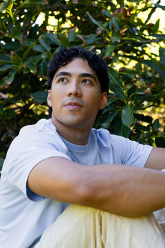 Asian man sitting, looking upward-left in garden, wearing light blue t-shirt among foliage