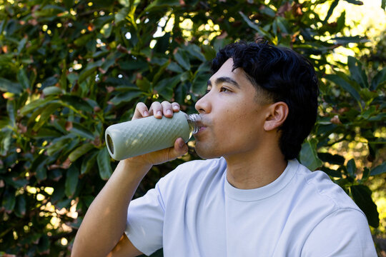 Man sitting among garden shrubs wearing light crewneck tee  and  drinking from textured reusable bot