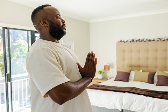Mid-adult African American man praying by bed near sliding door wearing wristband