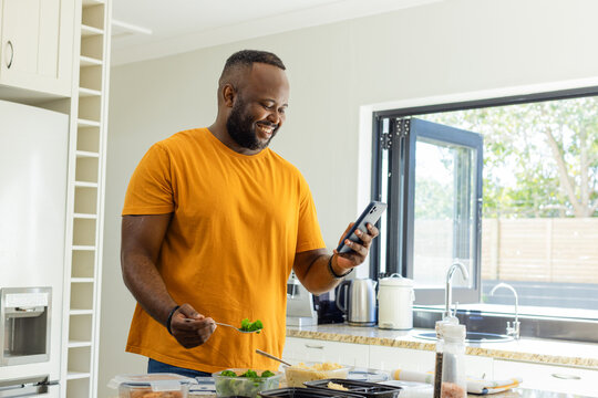 Mid adult man in orange tee packing meal containers while checking phone at kitchen counter
