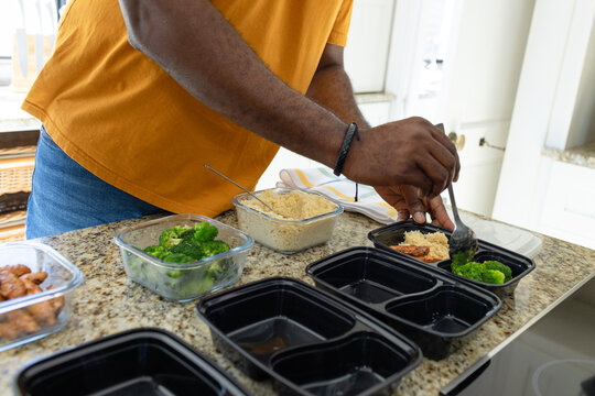 African man wearing mustard tee packing black meal-prep boxes with metal spoon at granite counter