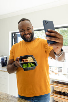 Middle-aged African man standing in home kitchen holding black divided container and angling phone
