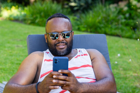 African American 30s man lounging on lounge chair in yard wearing striped tank holding blue phone