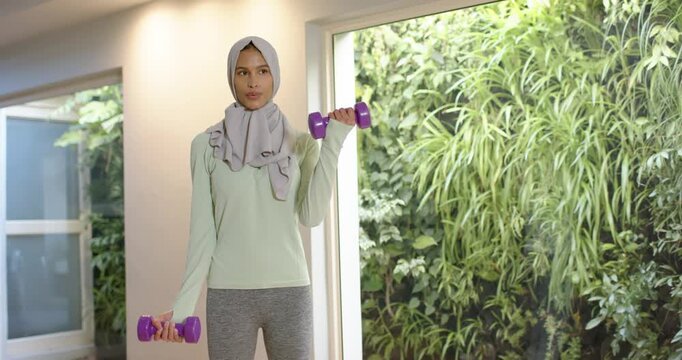 African American woman wearing hijab with purple weights home after cue curling arms by green wall
