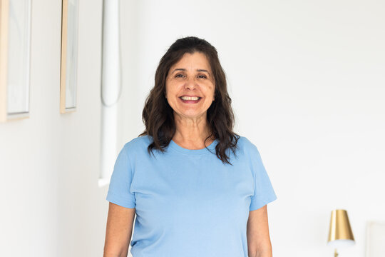 Woman wearing light blue T-shirt standing and smiling at home with framed art and lamp